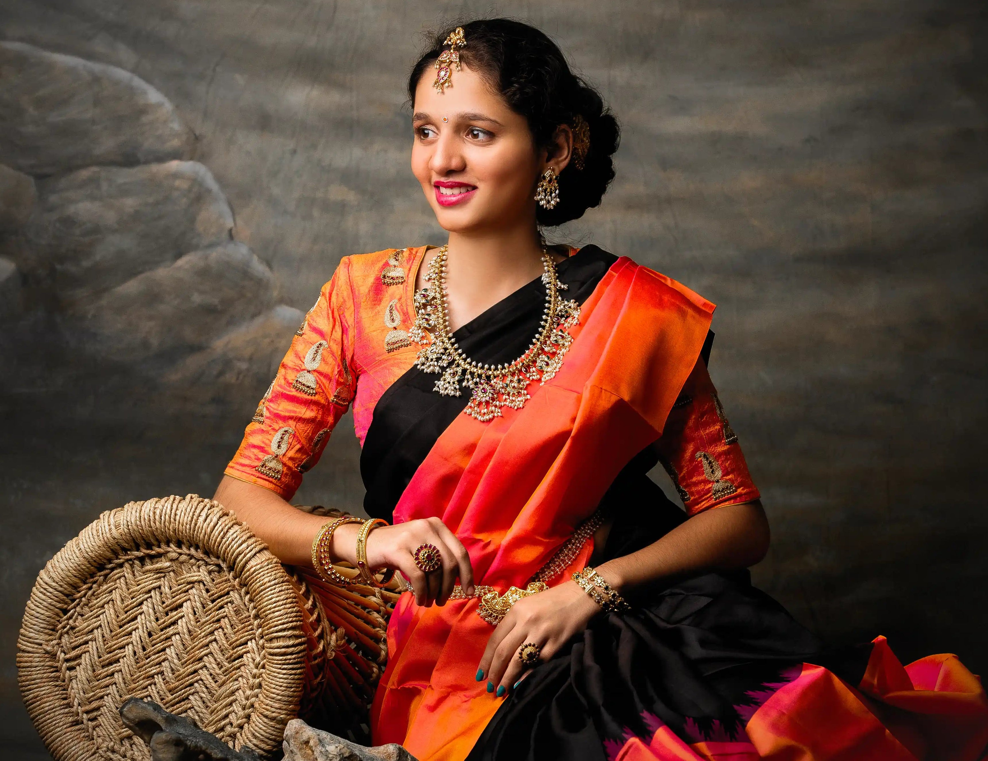Woman in traditional orange and black saree with jewelry against a textured dark background